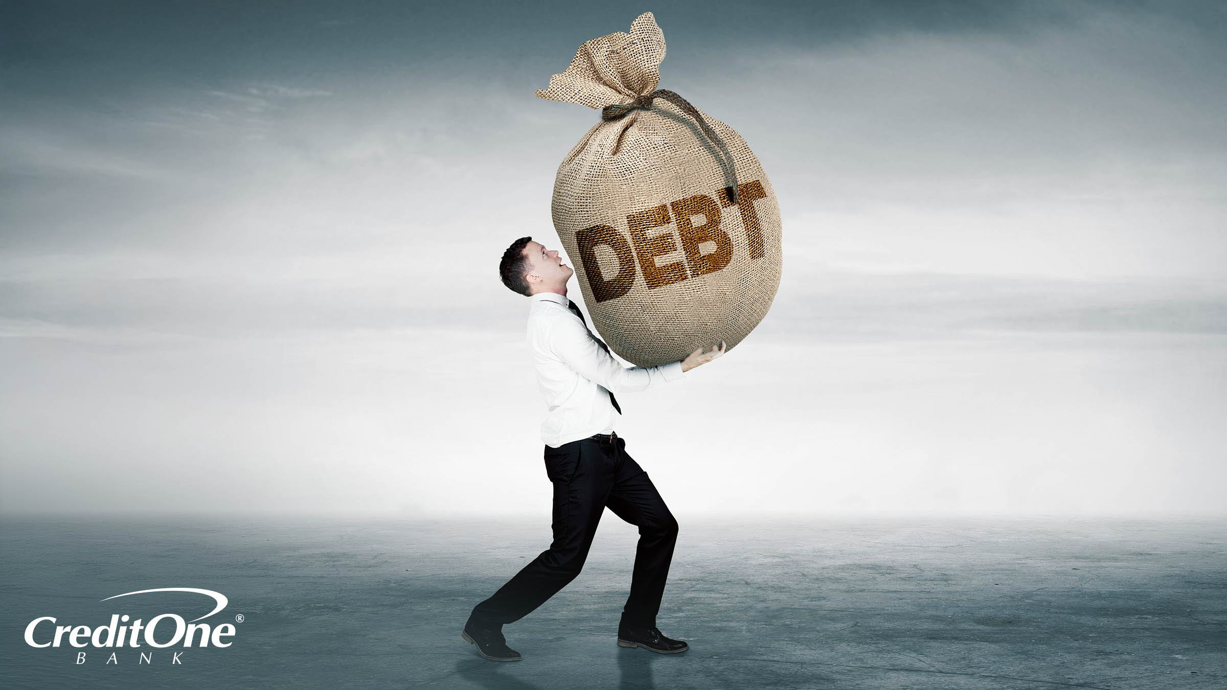 A young businessman struggles to carry a large burlap bag with the word “Debt” on it, representing the large burden of carrying debt like the average person.