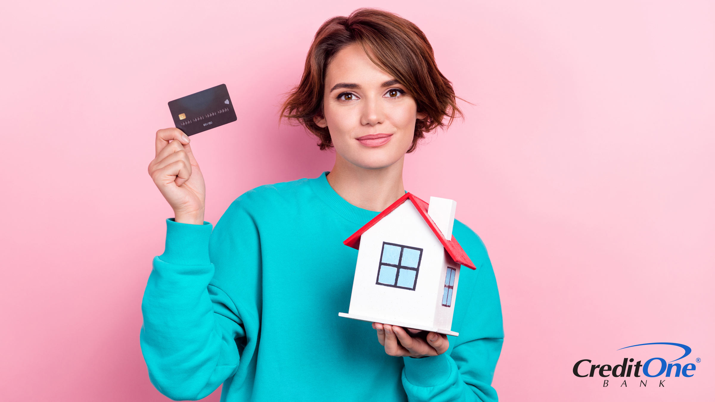 A young woman holds her credit card in one hand and a wooden model house in the other, representing the concept of paying a mortgage with a credit card.
