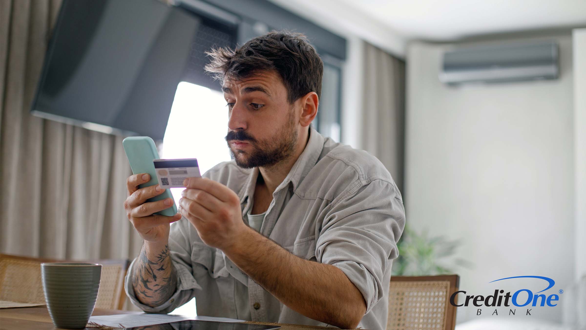 A man holds his phone in one hand and credit card in the other, looking into his phone with a surprised expression. Perhaps this is his first credit card and he narrowly avoided making a mistake.