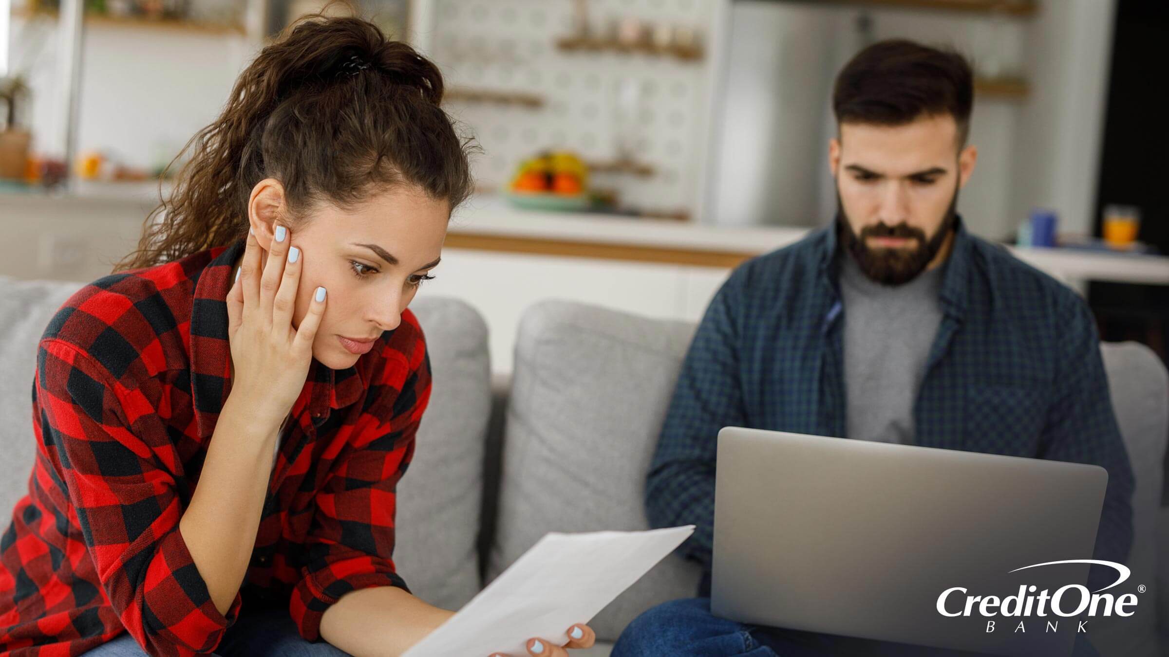 A couple sits on their sofa, the woman reading from a paper document, so the man can cross-check on his laptop. They may be in the process of disputing credit reporting errors.