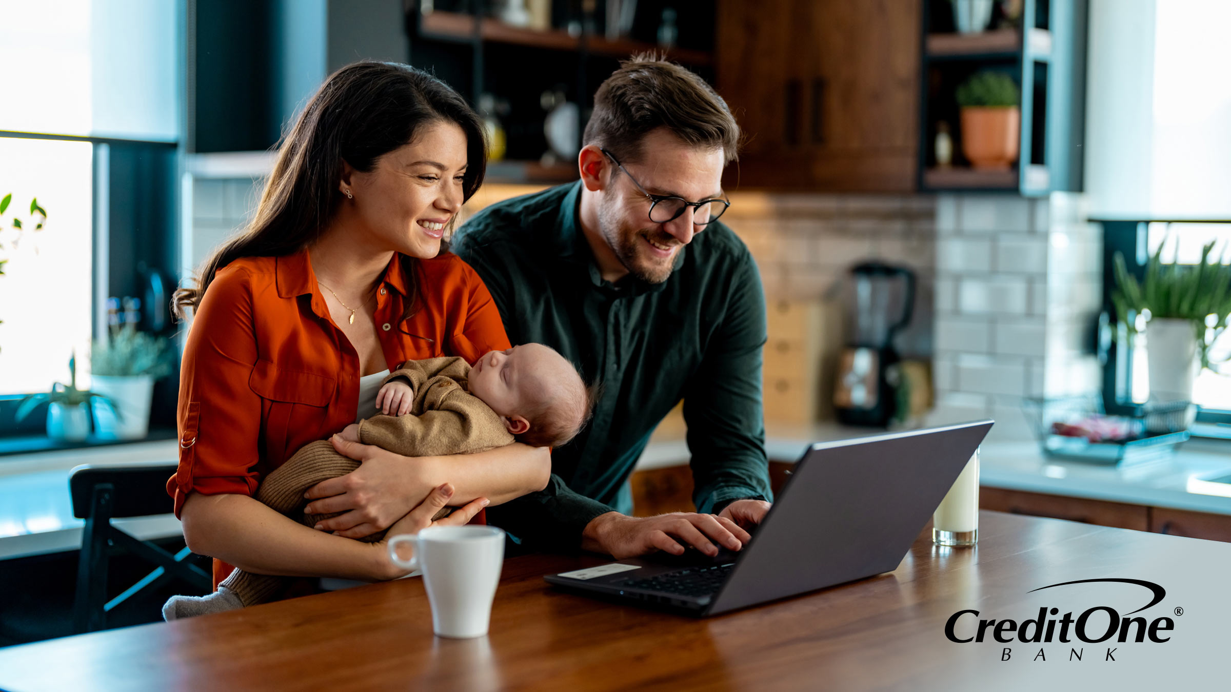 A couple reviews their finances on a laptop at the kitchen table, with the mother holding their newborn. They may be working on the family’s financial planning.