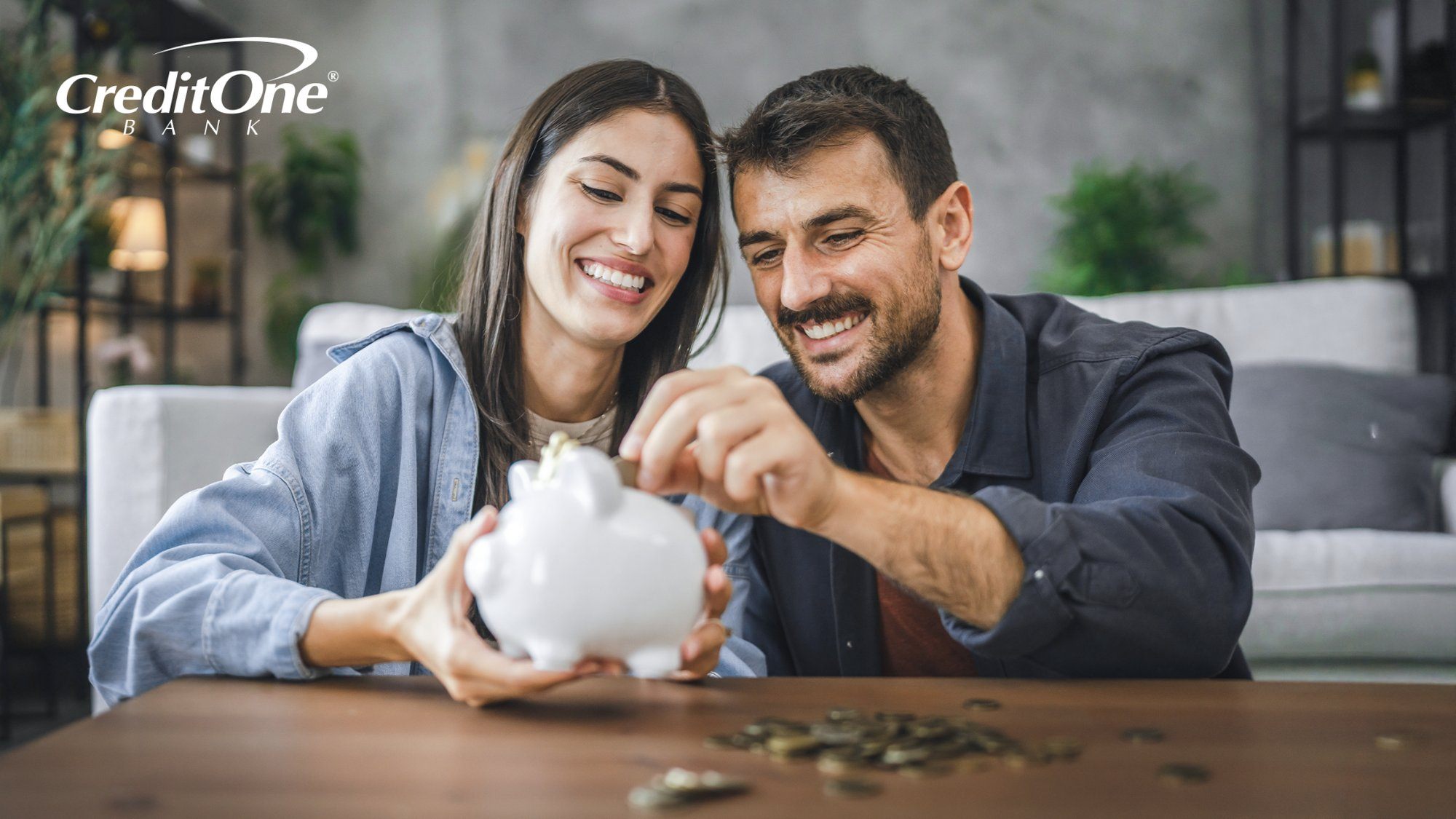 A smiling couple sits at their living room coffee table, placing coins from a scattered pile into a piggy bank. This represents learning how to invest and putting it into action.