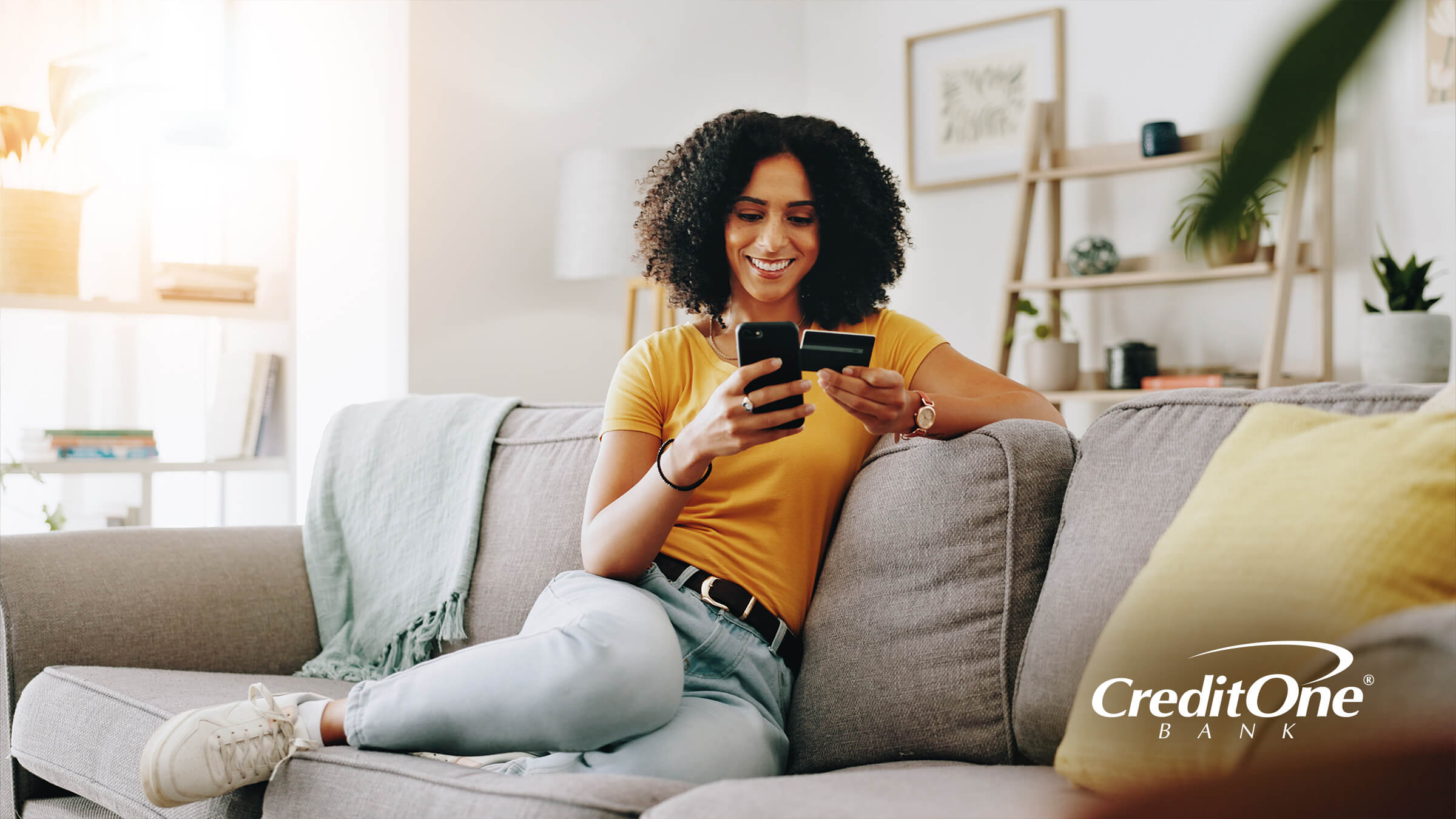 A woman rests on her sofa, holding up a credit card while happily looking at her phone. Perhaps she doesn’t use this card often and checked to see if the account is still open. 